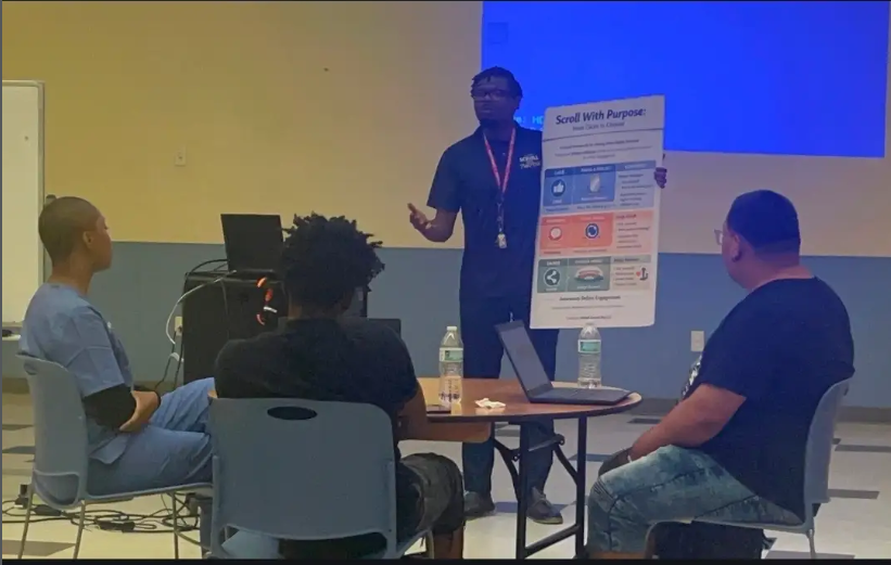 A facilitator presenting the Scroll With Purpose framework poster to a small group in a Philadelphia community program room